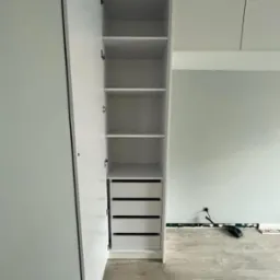 Otwarta, biała szafa wnękowa z półkami i czterema szufladami, widoczne okucia i fragment ściany w tle.