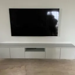 Szara szafka RTV na wymiar z telewizorem wiszącym nad nią, widoczne niedoskonałości na podłodze sugerują prace wykończeniowe.