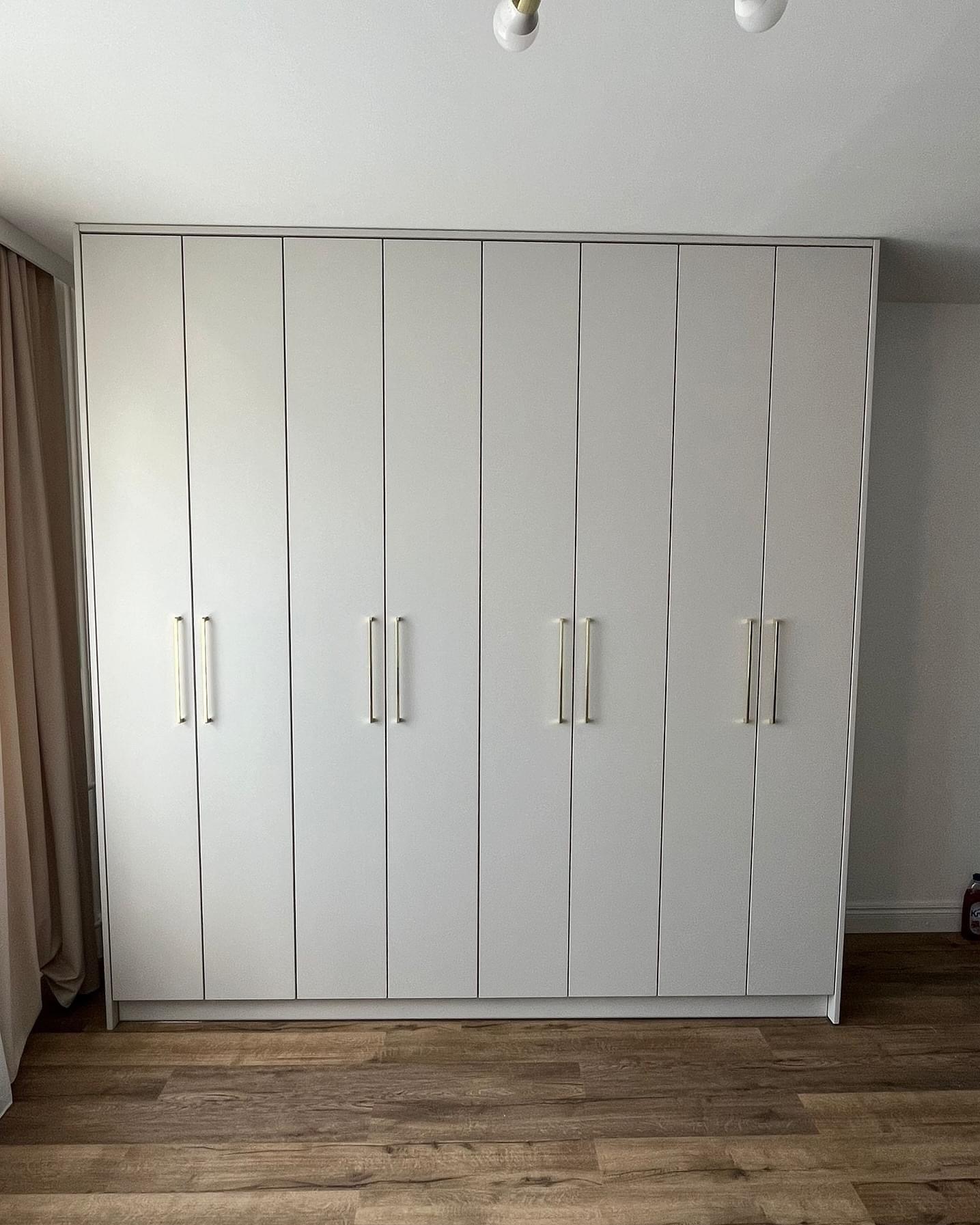 Duża, jasnoszara szafa wnękowa z siedmioma drzwiami i złotymi uchwytami, na drewnianej podłodze, z zasłoną po lewej stronie.