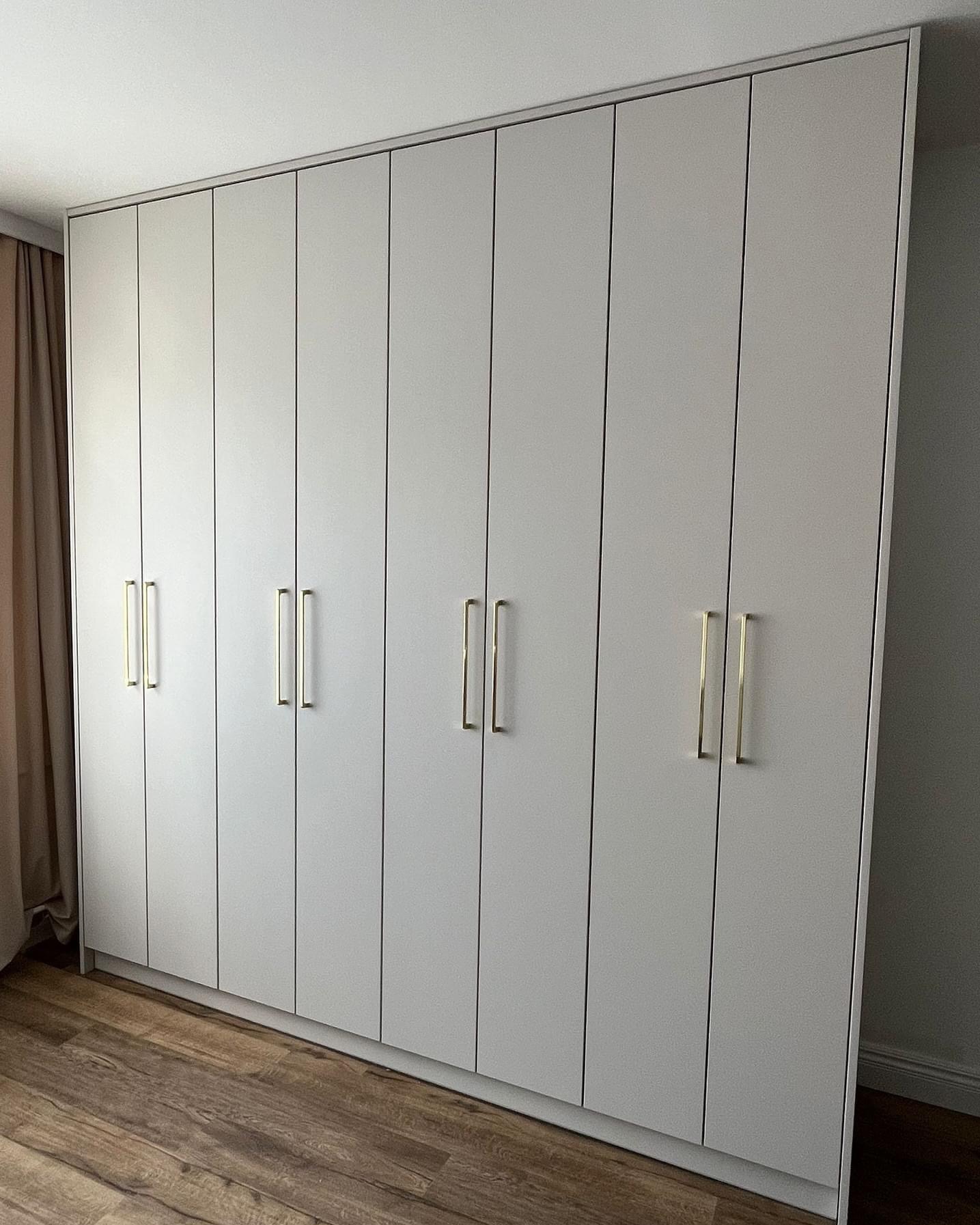 Biała szafa wnękowa z ośmioma drzwiami i złotymi, prostokątnymi uchwytami, widoczna na tle drewnianej podłogi i zasłony.