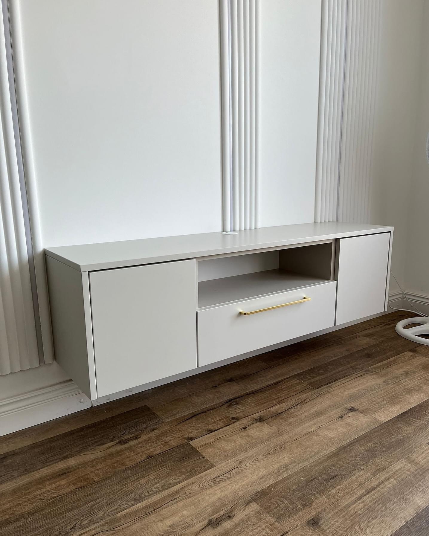 Biała szafka RTV wisząca na ścianie z dekoracyjnymi panelami, z szufladą z długim złotym uchwytem, na tle drewnianej podłogi