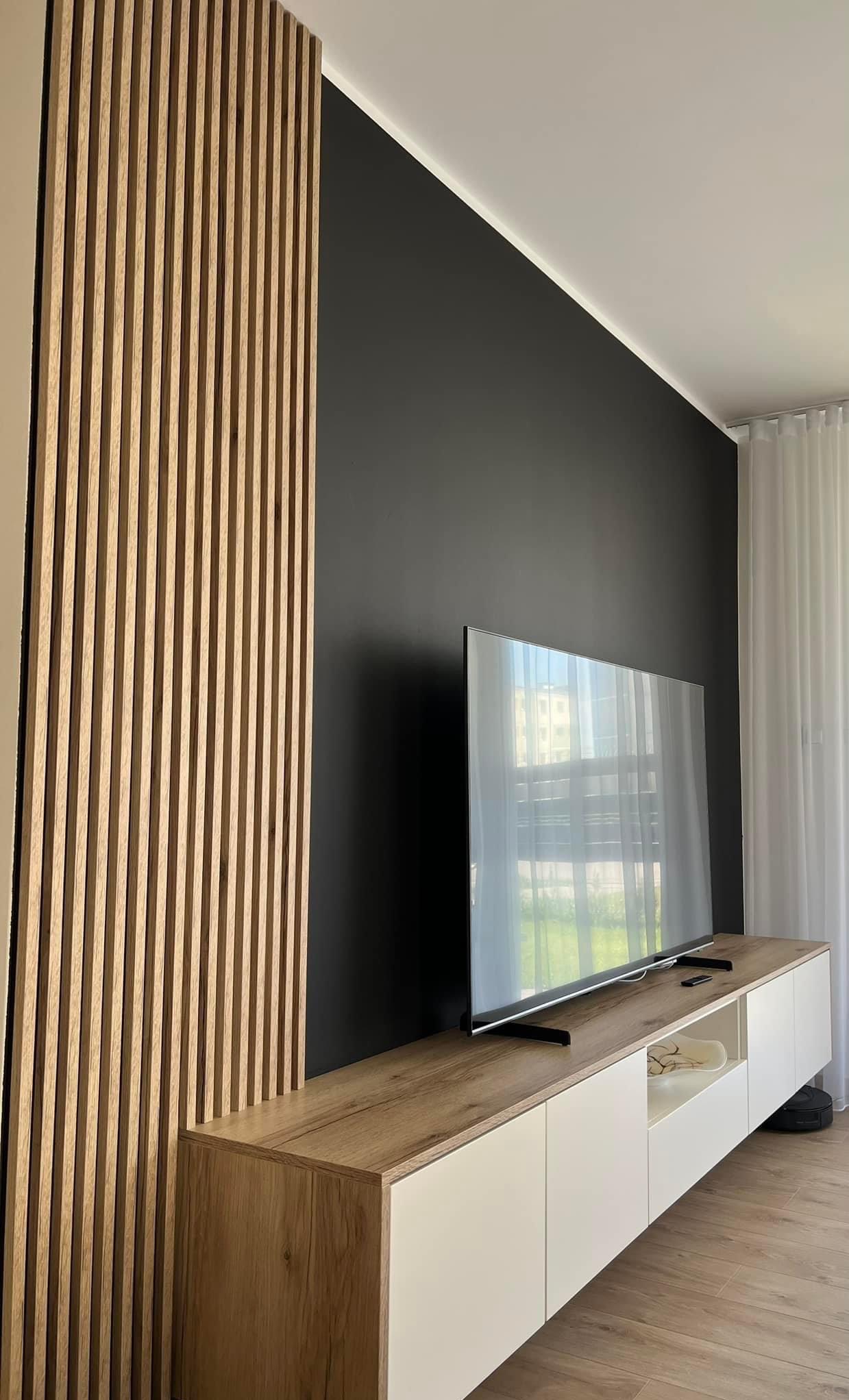 Nowoczesny salon z telewizorem umieszczonym na białej szafce RTV z drewnianym blatem, obok drewniana ściana z pionowych listew, czarna ściana w tle i białe zasłony w oknie.