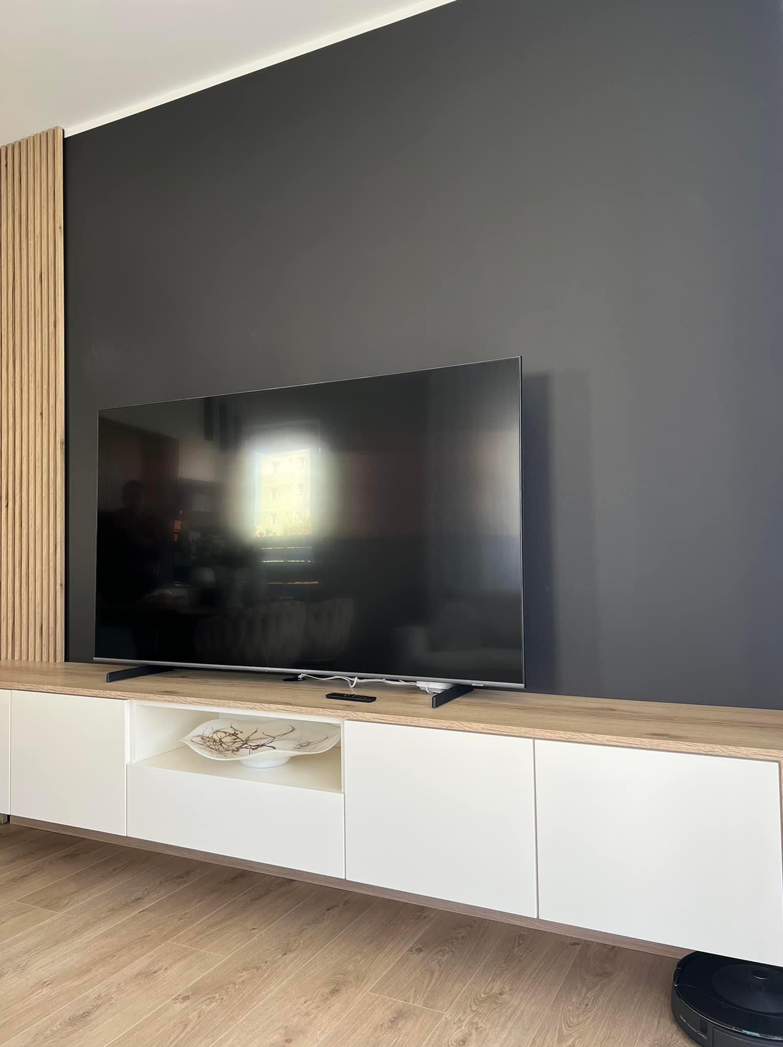 Nowoczesna szafka RTV z białymi frontami i drewnianym blatem, na tle ciemnej ściany z drewnianymi lamelami po lewej stronie, telewizor na szafce, robot sprzątający na podłodze.
