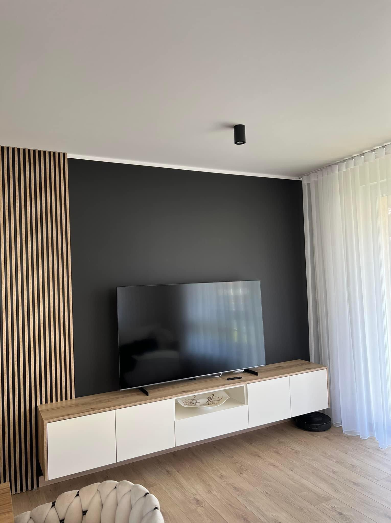 Nowoczesny salon z czarną ścianą, drewnianymi lamelami, białą szafką RTV i telewizorem; widoczny robot sprzątający i fragment fotela.