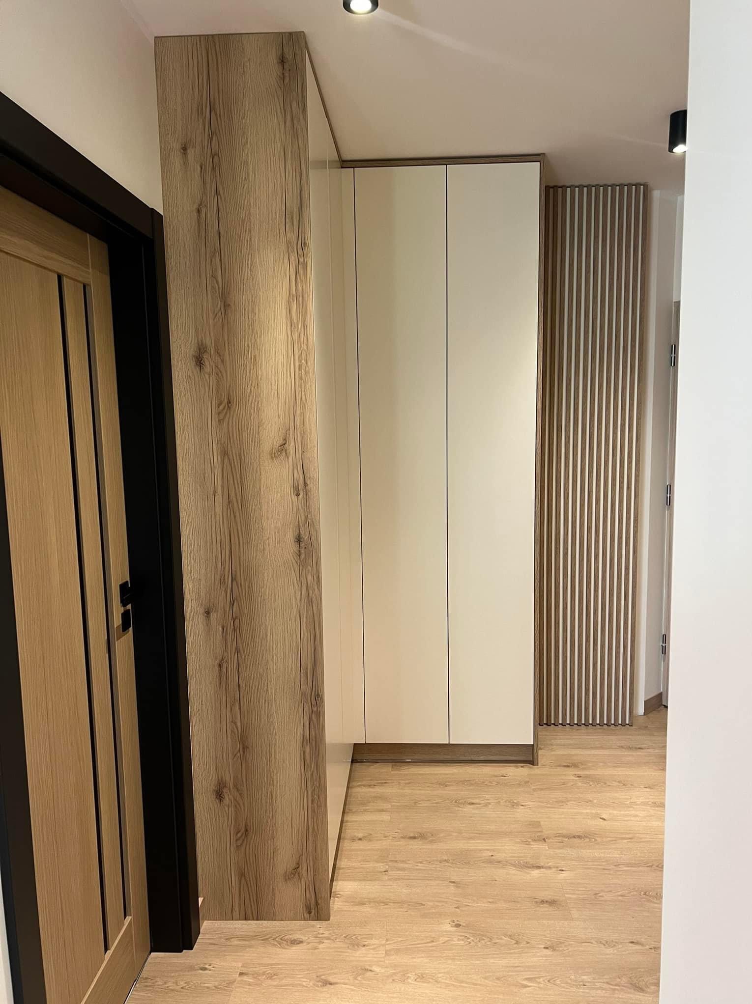 Nowoczesna zabudowa przedpokoju z szafą wnękową o białych frontach i drewnianym wykończeniu, dopasowana do koloru drzwi i podłogi, z dekoracyjnym panelem lamelowym w tle.