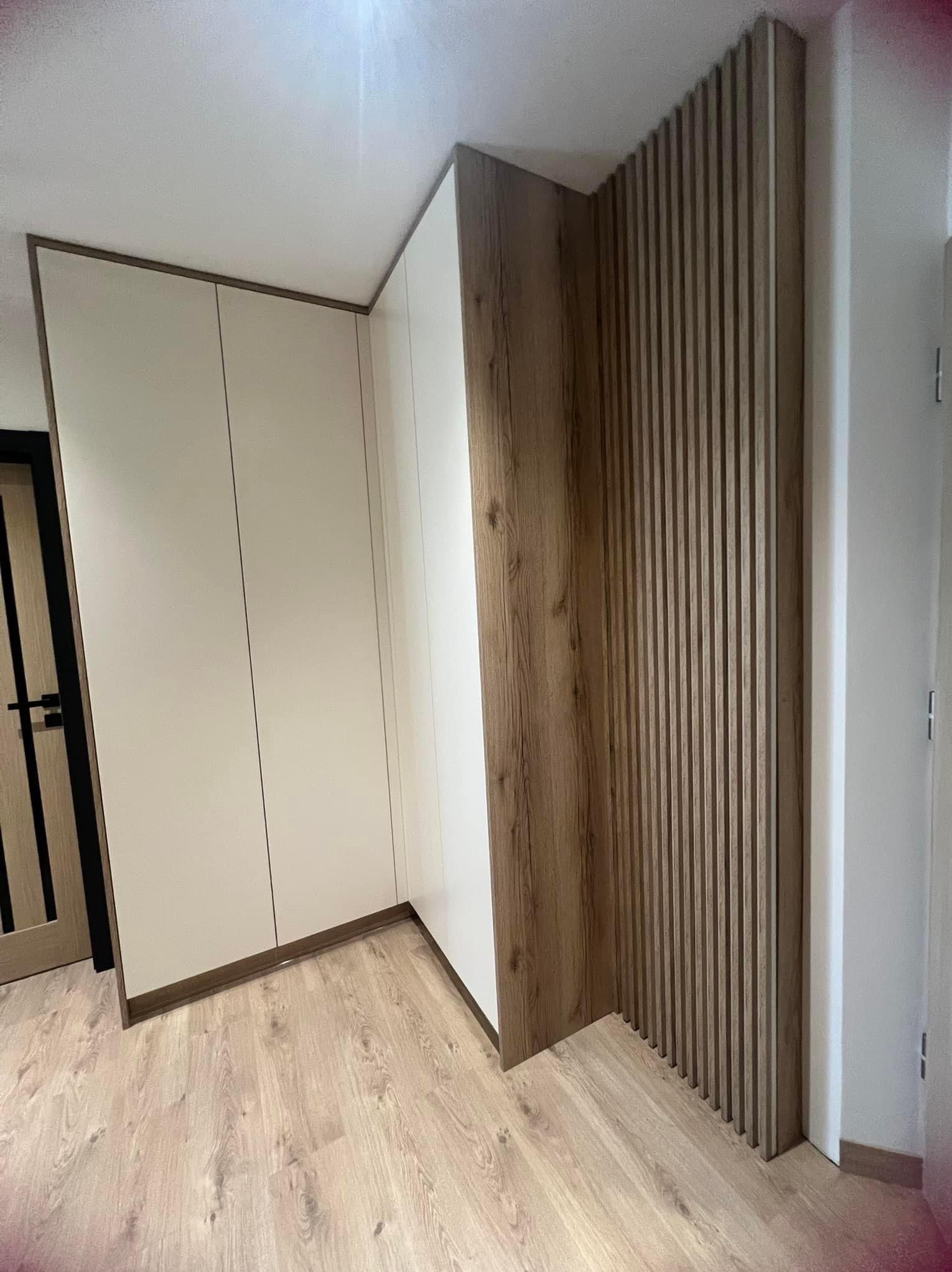 Biała szafa wnękowa z drewnianymi akcentami i dekoracyjną ścianką z drewnianych listewek w jasnym wnętrzu.