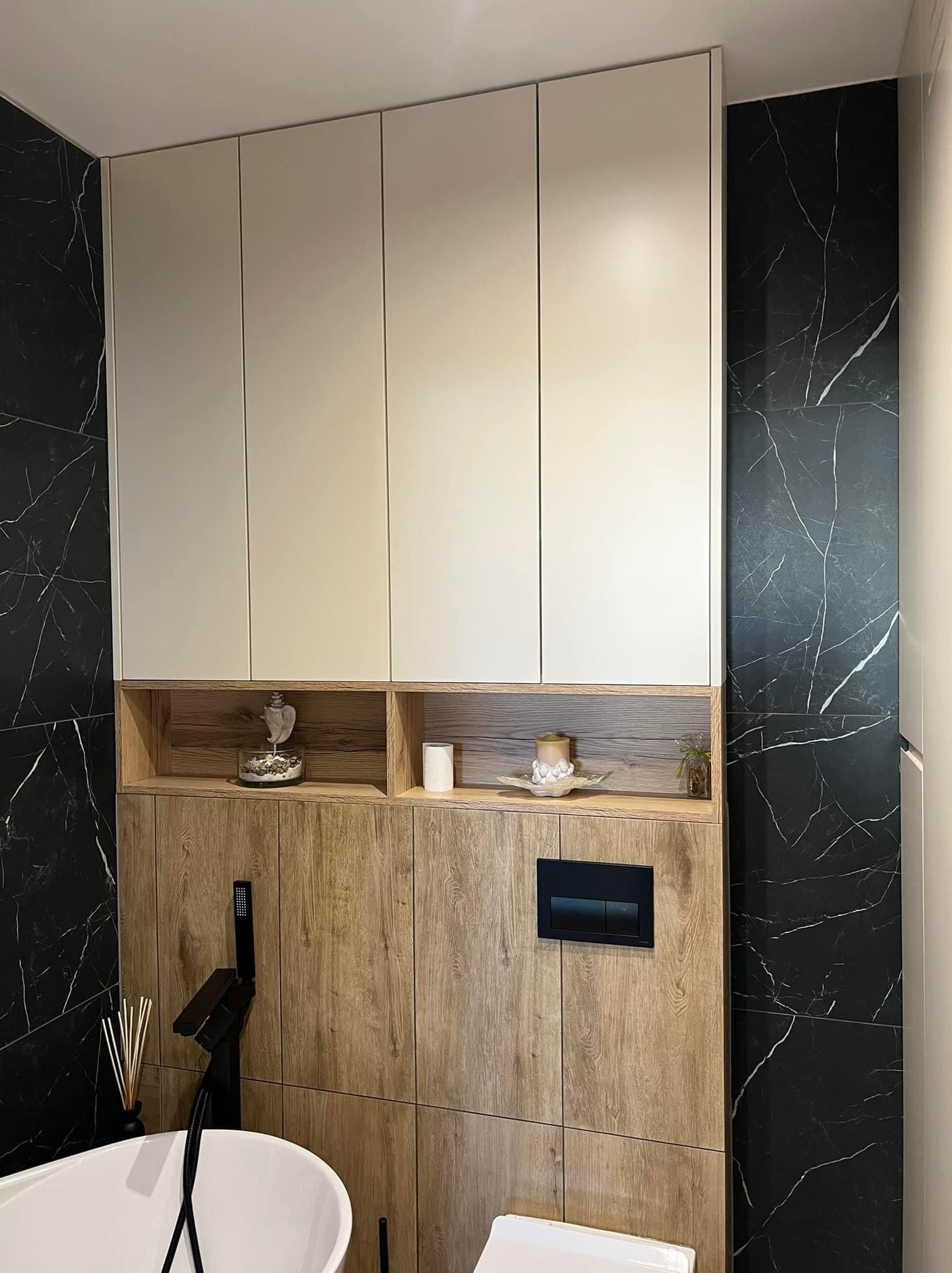 Nowoczesna łazienka z białymi szafkami wiszącymi, drewnianą zabudową, czarnym marmurem na ścianach i minimalistyczną armaturą.