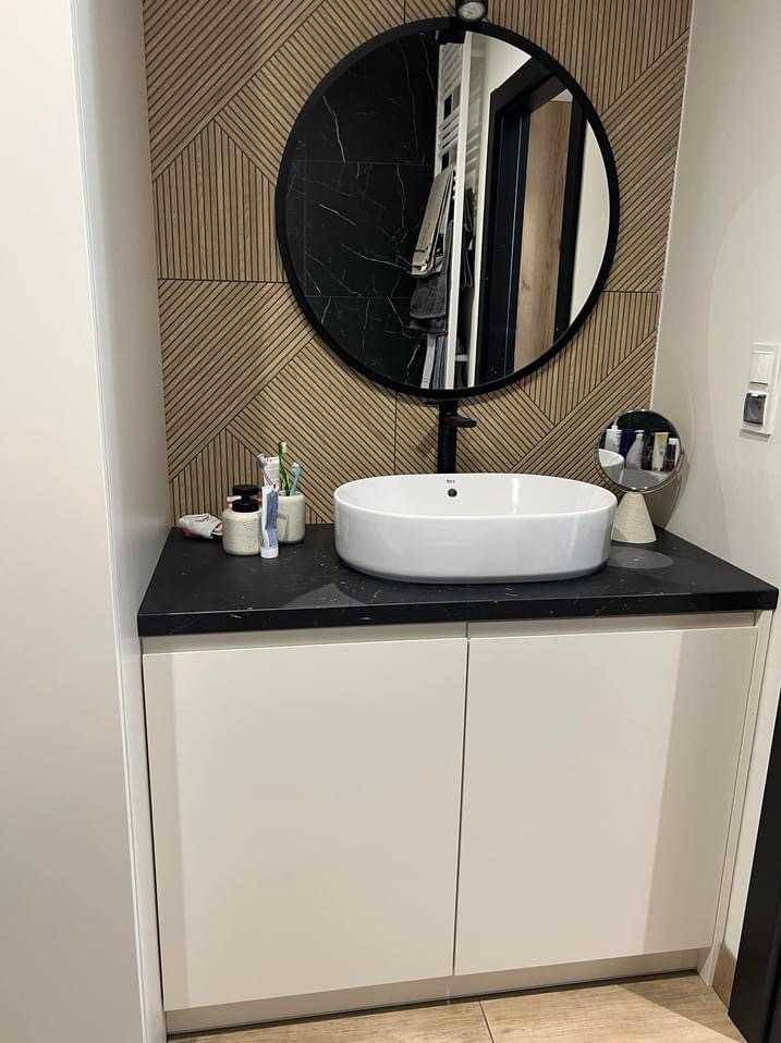 Nowoczesna łazienka z okrągłym lustrem, białą umywalką nablatową na czarnym blacie i szafką z białymi frontami. Na blacie kosmetyki i szczoteczki do zębów. W tle ściana z drewnianymi panelami.