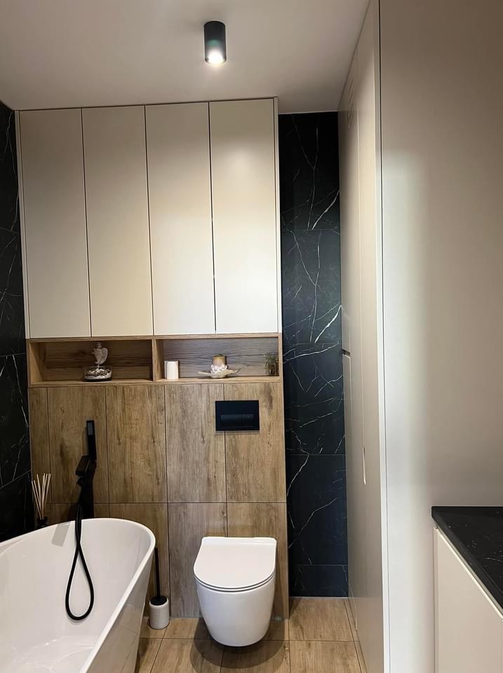 Nowoczesna łazienka z wanną wolnostojącą i zabudową meblową w Elblągu, z białymi szafkami górnymi i drewnopodobnymi panelami dolnymi, uzupełniona czarnym marmurem na ścianach.