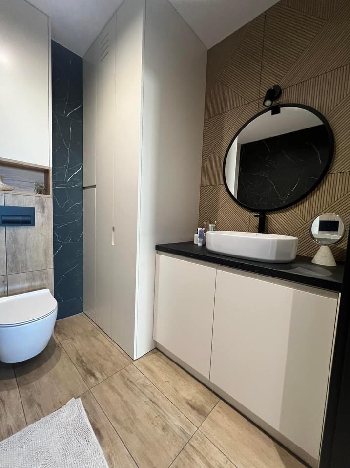 Nowoczesna łazienka z drewnopodobną podłogą, szafką pod umywalką z czarnym blatem, owalną umywalką nablatową, okrągłym lustrem z czarną ramą i wysoką szafą w zabudowie.