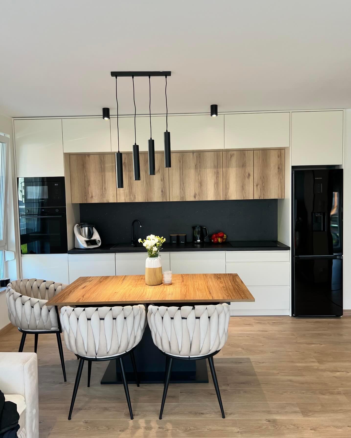 Nowoczesna kuchnia z białymi i drewnianymi frontami, czarnym blatem, wiszącymi lampami i stołem z krzesłami o plecionych oparciach.