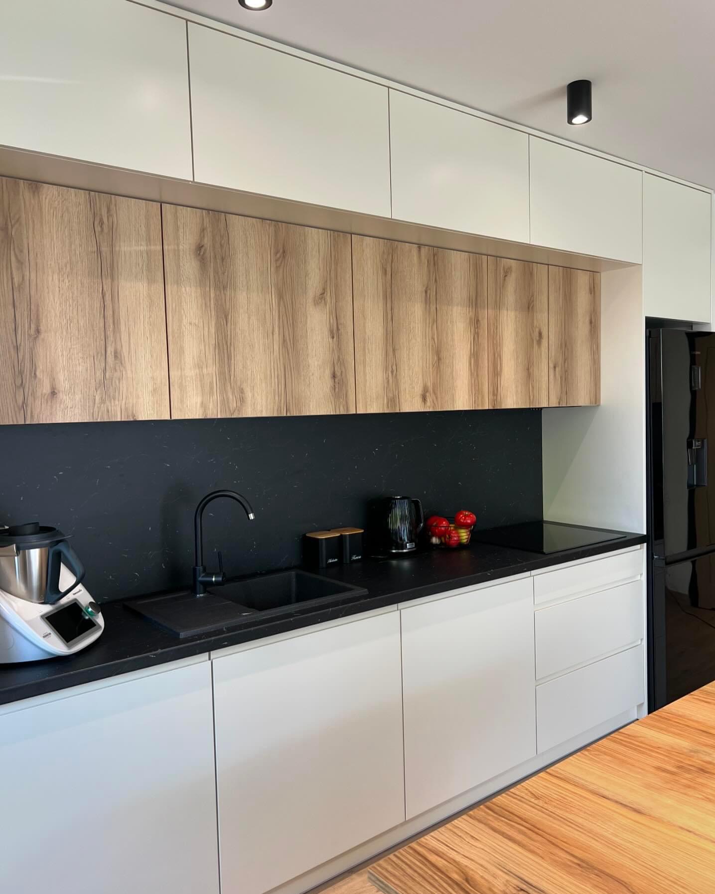Nowoczesna kuchnia z białymi i drewnopodobnymi szafkami, czarnym blatem i backsplash'em, zintegrowanym zlewem i czarną armaturą, oraz Thermomixem po lewej stronie.