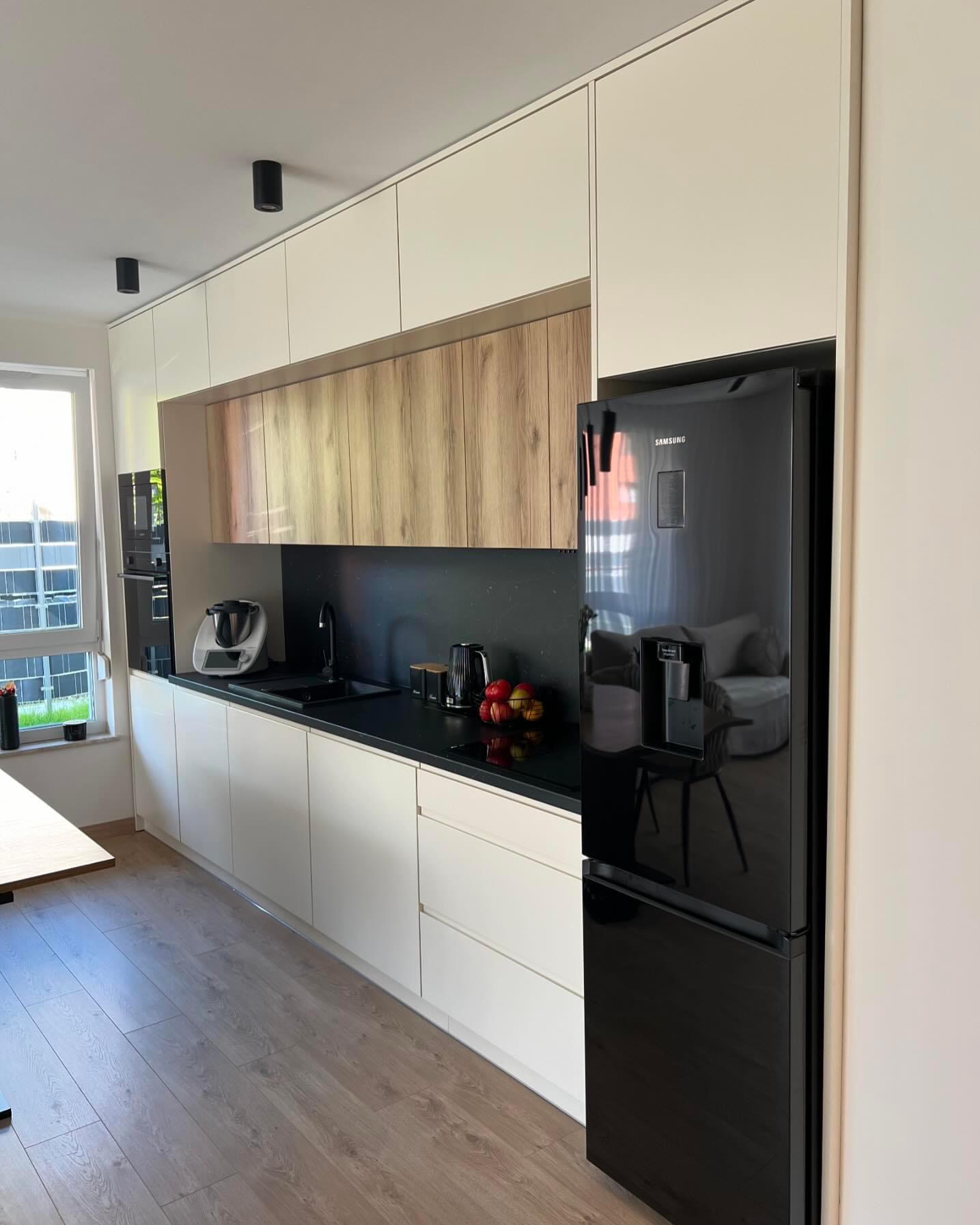 Nowoczesna kuchnia z białymi szafkami dolnymi i górnymi, drewnopodobnymi szafkami wiszącymi, czarnym blatem i czarną lodówką Samsung z dystrybutorem wody.