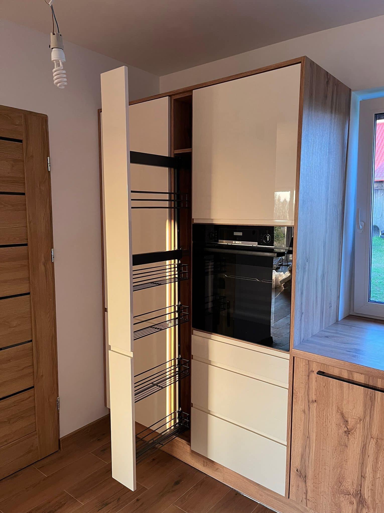 Nowoczesna zabudowa kuchenna z otwartym, wysuwanym systemem cargo, jasnymi frontami i wykończeniem w kolorze drewna, z widocznym piekarnikiem w zabudowie.