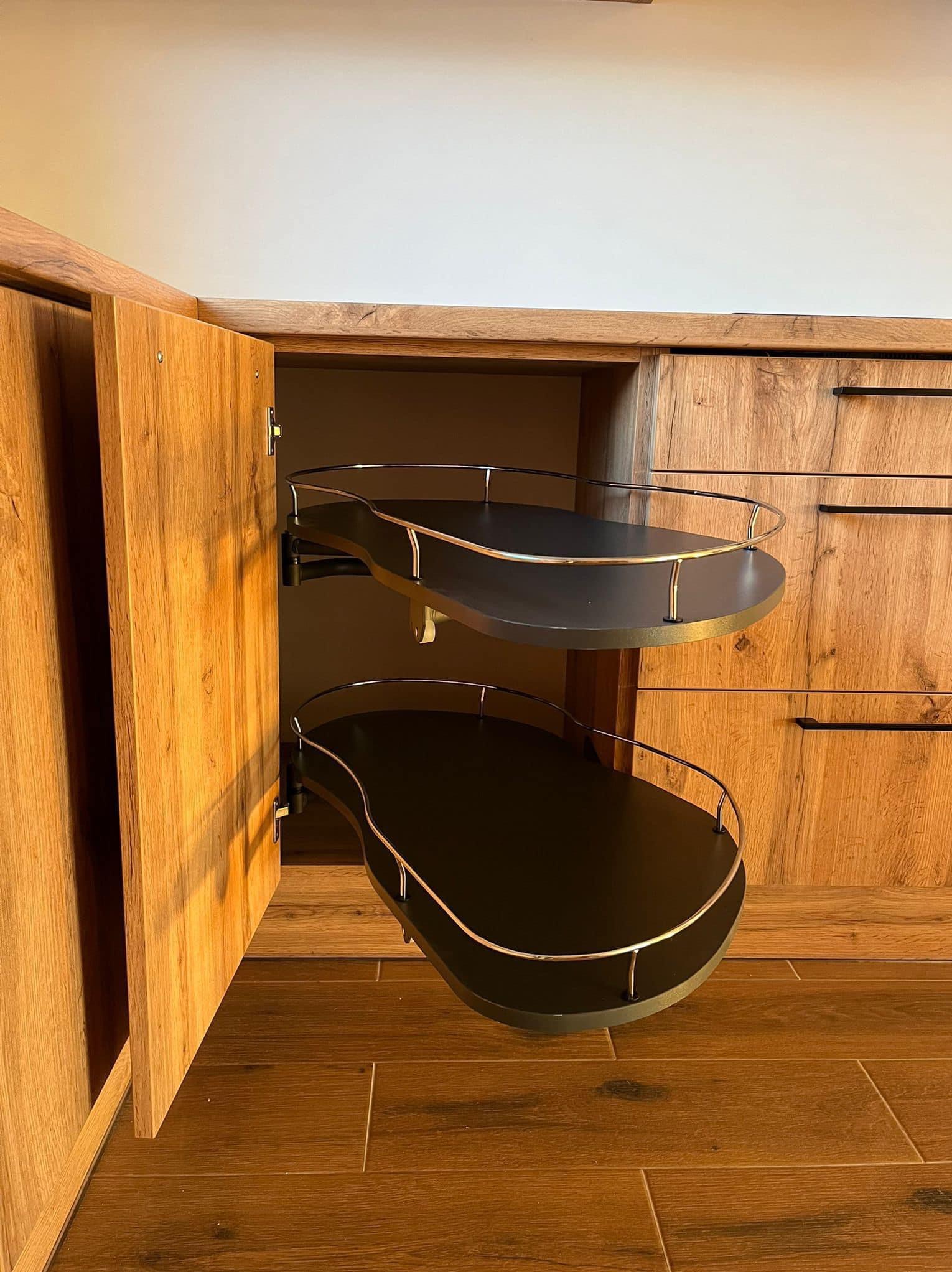 Wnętrze szafki narożnej z drewnianymi frontami, wyposażonej w dwupoziomowy system półek obrotowych z metalowymi relingami, widoczne na tle drewnianej podłogi i innych elementów zabudowy kuchennej.