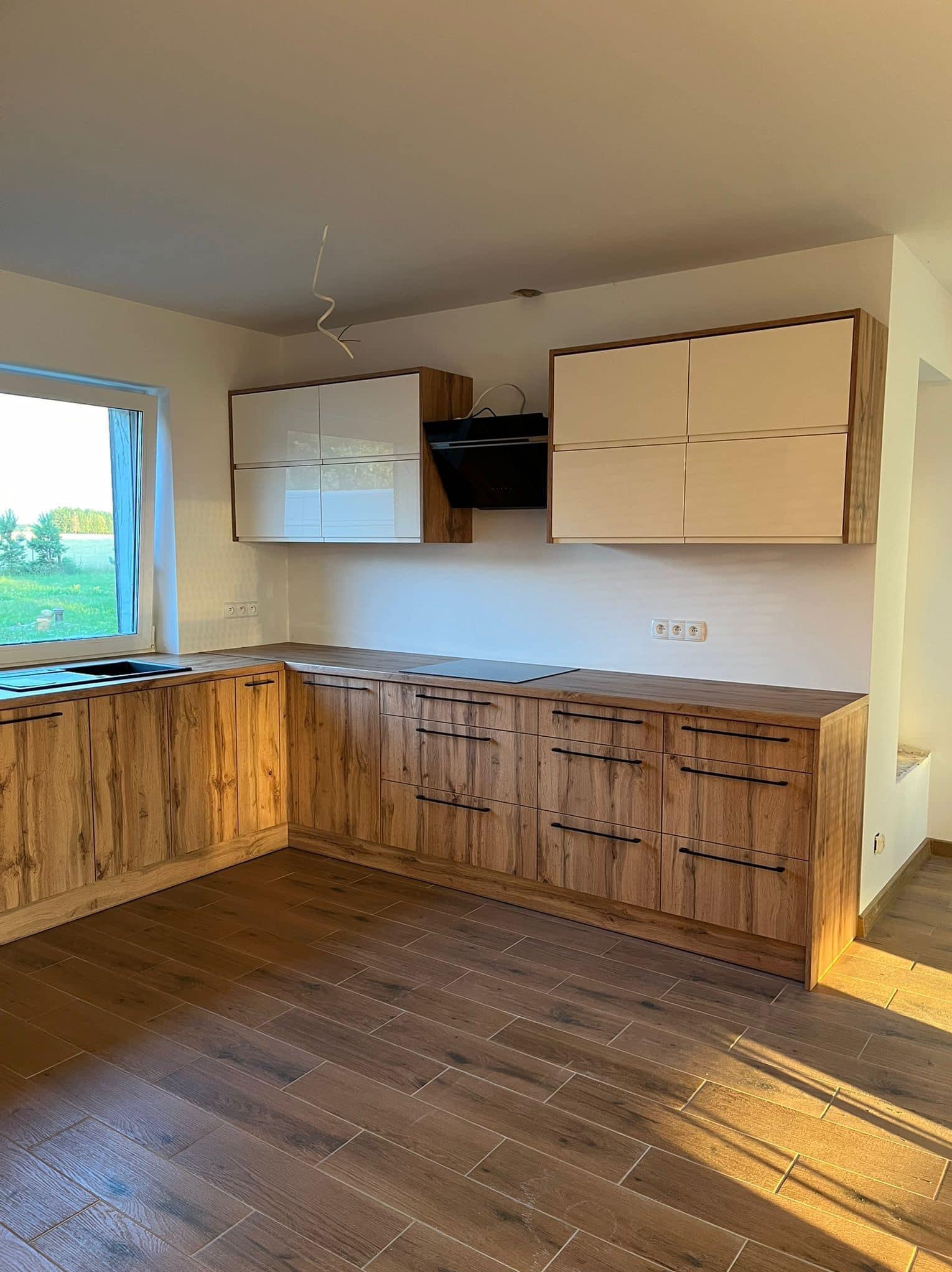 Nowoczesna kuchnia z drewnianymi frontami szafek dolnych i białymi szafek górnych, czarny okap, drewniana podłoga, widok na zewnątrz przez okno.