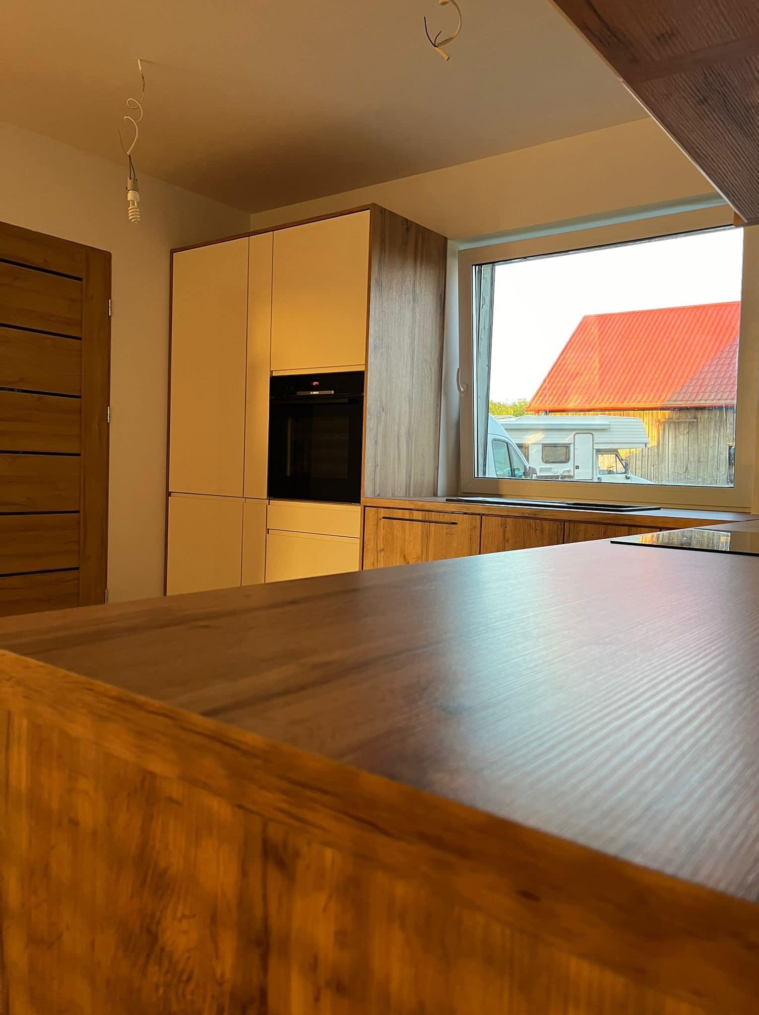 Fragment nowej kuchni z drewnianymi i białymi frontami, widoczny piekarnik w zabudowie, blat roboczy i okno z widokiem na dom z pomarańczowym dachem oraz kamper.