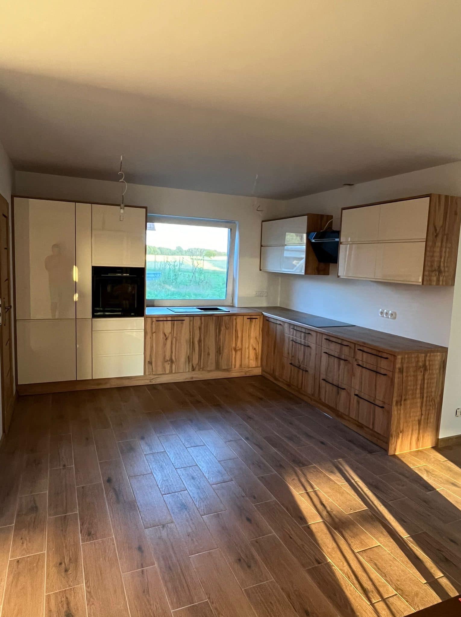 Nowoczesna kuchnia z drewnianymi frontami i jasnymi szafkami, widok na zewnątrz przez okno, brązowe płytki podłogowe w promieniach słońca.