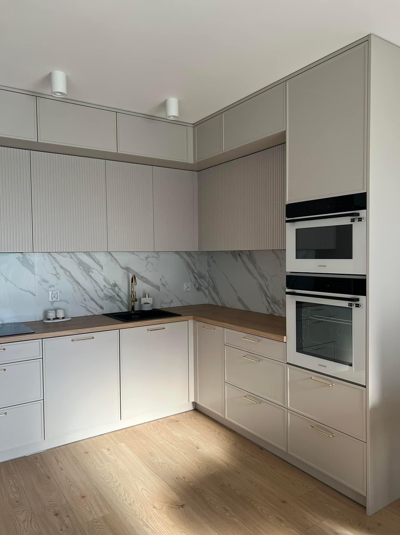 Jasna kuchnia z frezowanymi frontami górnych szafek, marmurowym backsplashiem, złotą armaturą i wbudowanymi urządzeniami AGD marki Samsung.