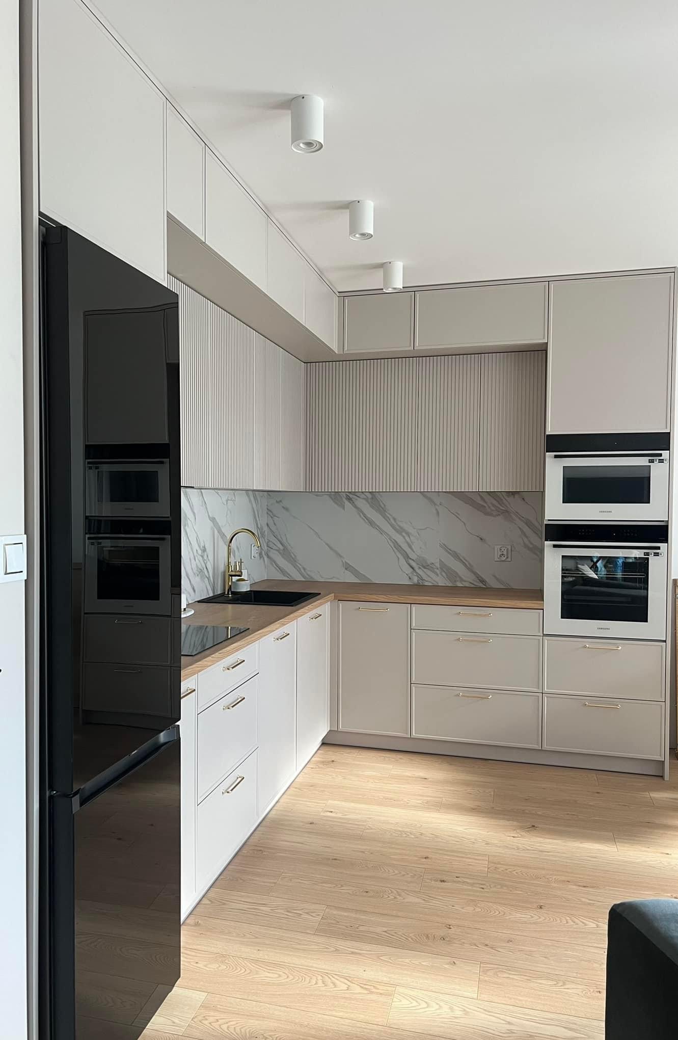 Jasna kuchnia z frezowanymi frontami szafek wiszących, marmurowym panelem nad blatem, złotą armaturą i zabudowanymi sprzętami AGD.