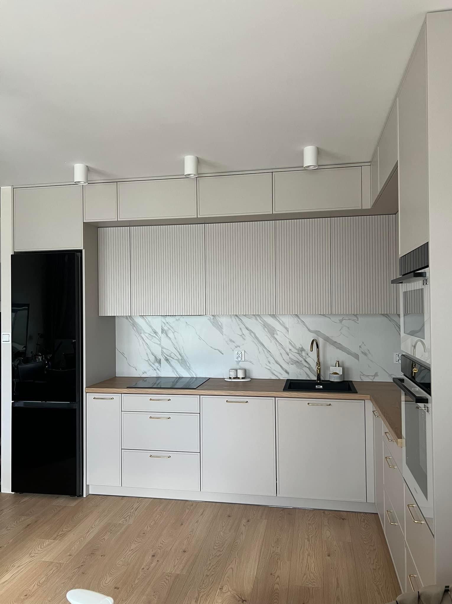 Jasna kuchnia z frezowanymi frontami szafek górnych, marmurowym backsplashiem i złotą armaturą, czarna lodówka po lewej stronie.