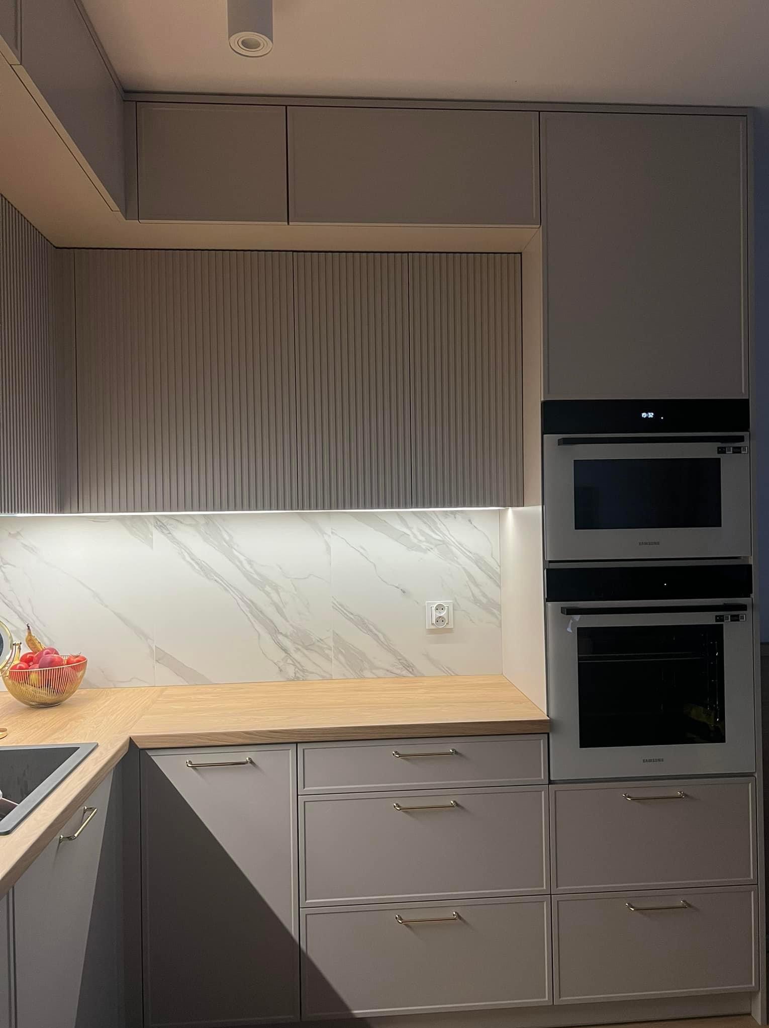 Nowoczesna kuchnia w odcieniach szarości z drewnianym blatem, marmurowymi płytkami na ścianie i zabudowanymi piekarnikami Samsung.