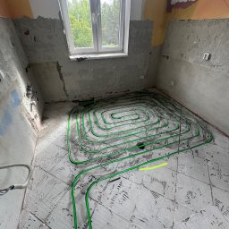Cempa Instalacje Maciej Cempa - Układanie zielonych rur ogrzewania podłogowego w surowym wnętrzu. Widok na okno i fragment instalacji na ścianie.