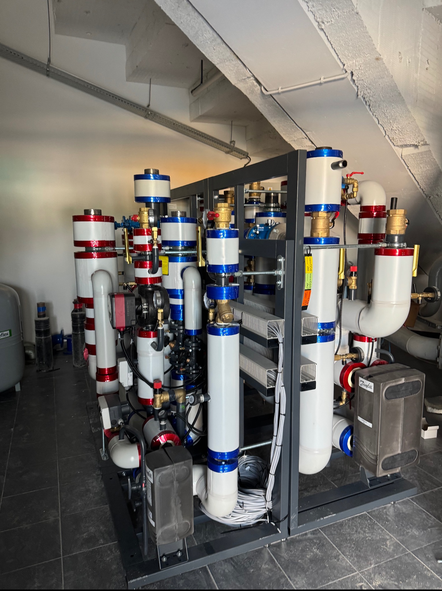 Złożona instalacja grzewcza z licznymi rurami, zaworami i urządzeniami w piwnicy, pod skosnym sufitem, z widocznymi elementami izolacji termicznej.