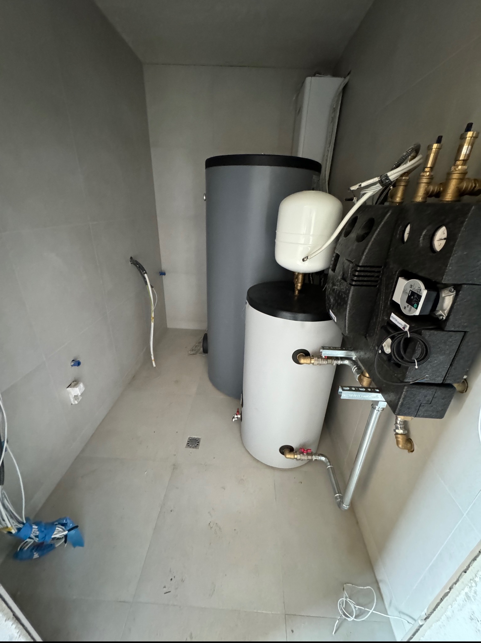 Szary i biały bojler w rogu pomieszczenia z szarymi płytkami. Widoczne rury i kable. Ujęcie z góry, ukazujące instalację w trakcie montażu.