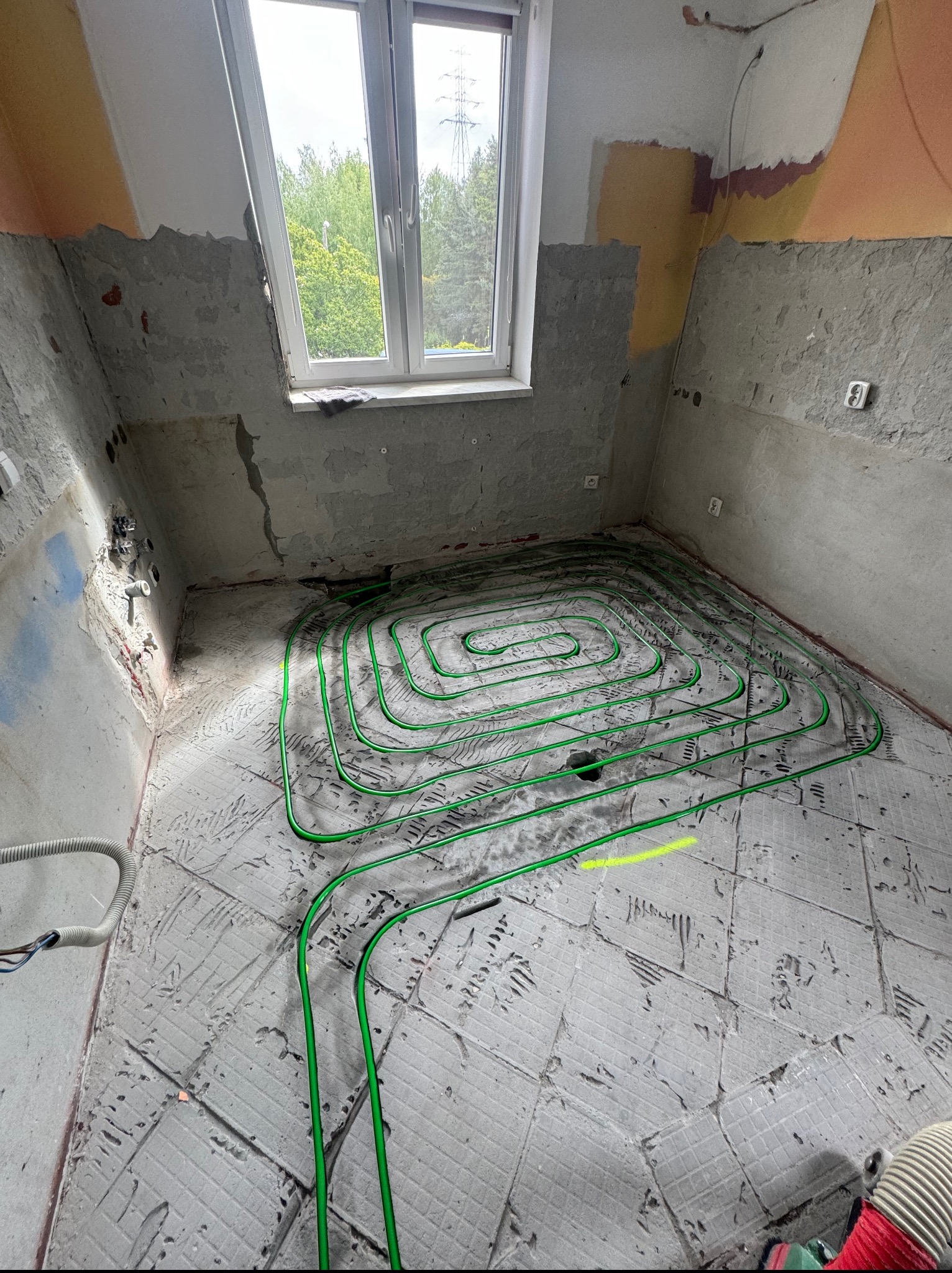 Układanie zielonych rur ogrzewania podłogowego w surowym wnętrzu. Widok na okno i fragment instalacji na ścianie.