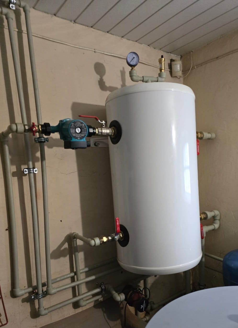 Biały zbiornik akumulacyjny ciepłej wody z zieloną pompą obiegową i manometrem, rury instalacji grzewczej w piwnicy.