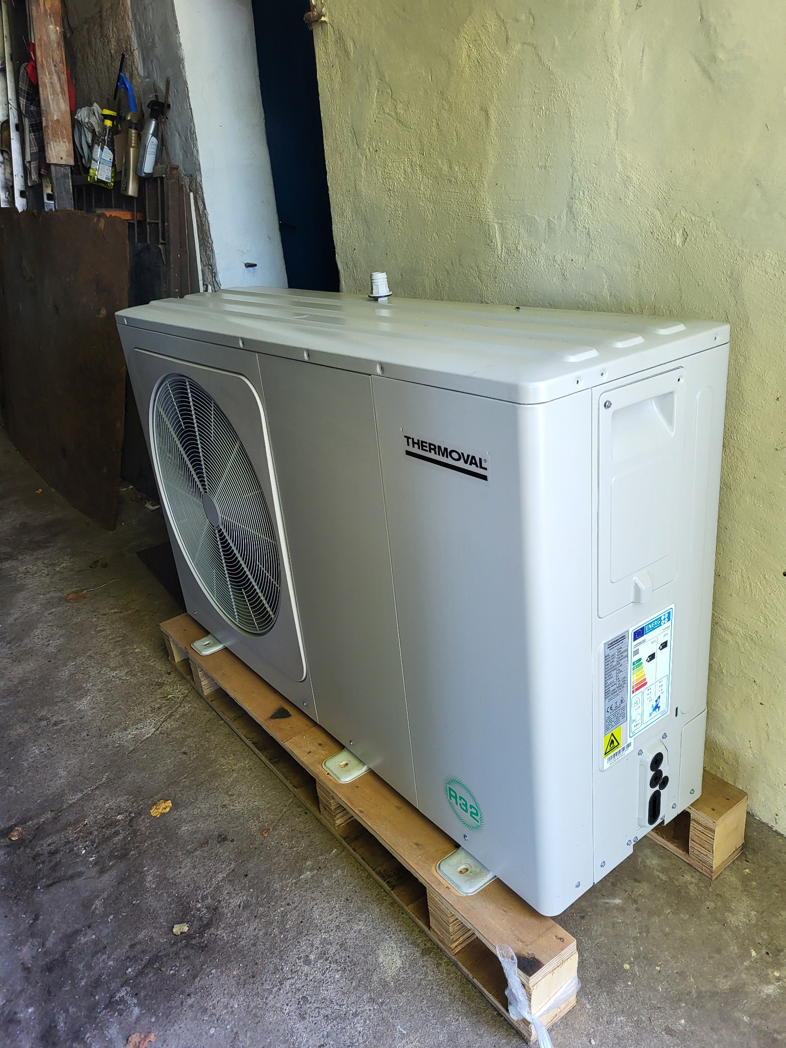 Szara pompa ciepła marki Thermoval umieszczona na drewnianej palecie wewnątrz budynku, widoczna tabliczka znamionowa i wentylator.