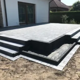 Nowoczesny taras z szarej kostki brukowej, schody z trzech stopni, obrzeża z ciemnoszarej kostki, widok na ogród i dom.