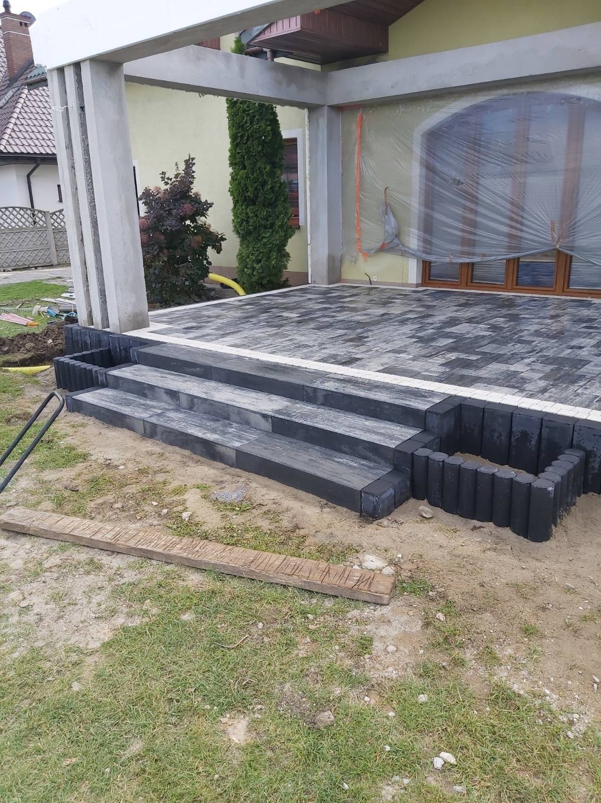 Nowo ułożony taras z kostki brukowej w odcieniach szarości, z trzema schodami prowadzącymi do zadaszonego patio. Widoczne elementy wykończeniowe z okrągłych, czarnych elementów betonowych. Dom...