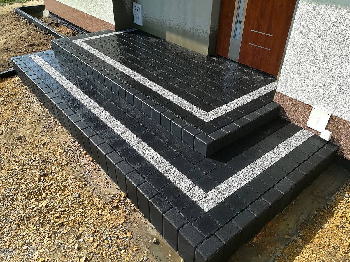 Schody wejściowe wyłożone czarną kostką brukową z dekoracyjnymi pasami z jasnej kostki granitowej, świeżo po deszczu, widoczne połączenie z elewacją budynku.