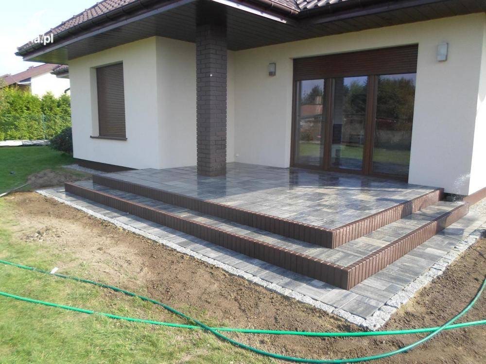 Wykończony taras z kostki brukowej w odcieniach szarości i brązu, z trzema stopniami obłożonymi brązowym obrzeżem, przylegający do budynku z brązową stolarką okienną i ceglanym filarem.