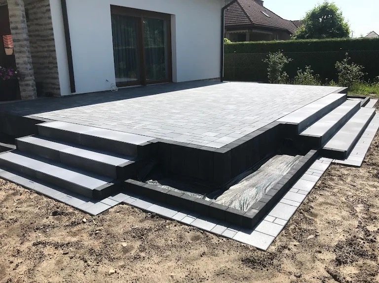 Nowoczesny taras z szarej kostki brukowej, schody z trzech stopni, obrzeża z ciemnoszarej kostki, widok na ogród i dom.