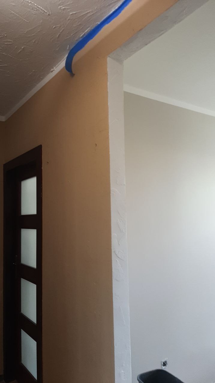 Przygotowanie do malowania: zabezpieczona taśmą malarską krawędź sufitu, ściana w kolorze ciepłego beżu, widoczny fragment drzwi z szybami oraz pomalowana na biało ściana w sąsiednim pomieszczeniu.