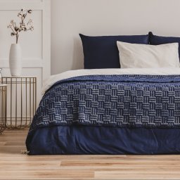 Odlotowy Home Staging - Elegancka sypialnia z łóżkiem w granatowej kolorystyce, wzorzysta narzuta, dwie metalowe szafki nocne z książkami i wazonem z bawełną, drewniana podłoga.
