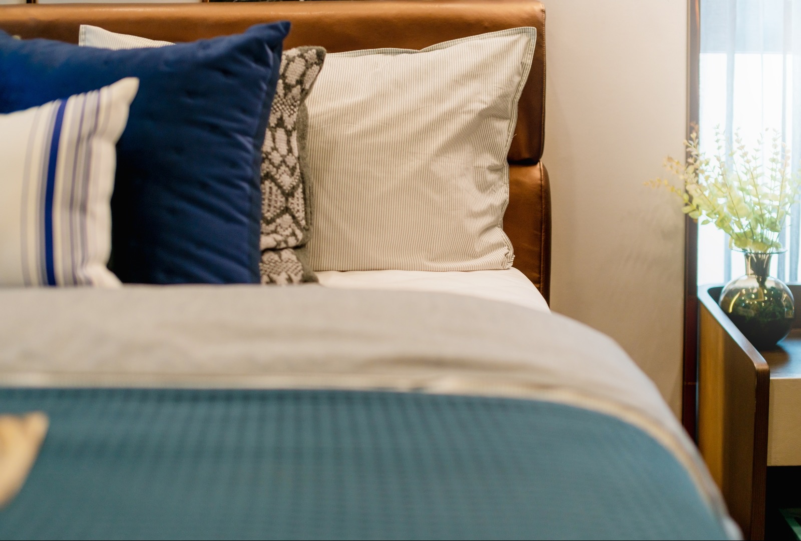 Detale sypialni: łóżko z brązowym skórzanym zagłówkiem, dekoracyjne poduszki w odcieniach granatu, szarości i bieli, oraz wazon z zielonymi gałązkami na drewnianym stoliku nocnym przy oknie.