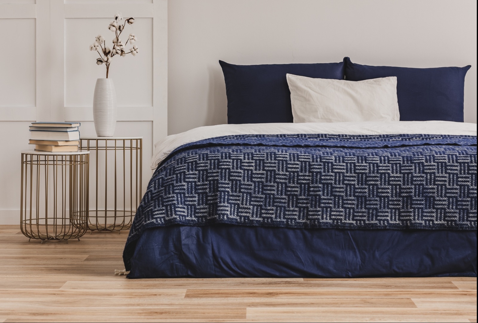 Elegancka sypialnia z łóżkiem w granatowej kolorystyce, wzorzysta narzuta, dwie metalowe szafki nocne z książkami i wazonem z bawełną, drewniana podłoga.
