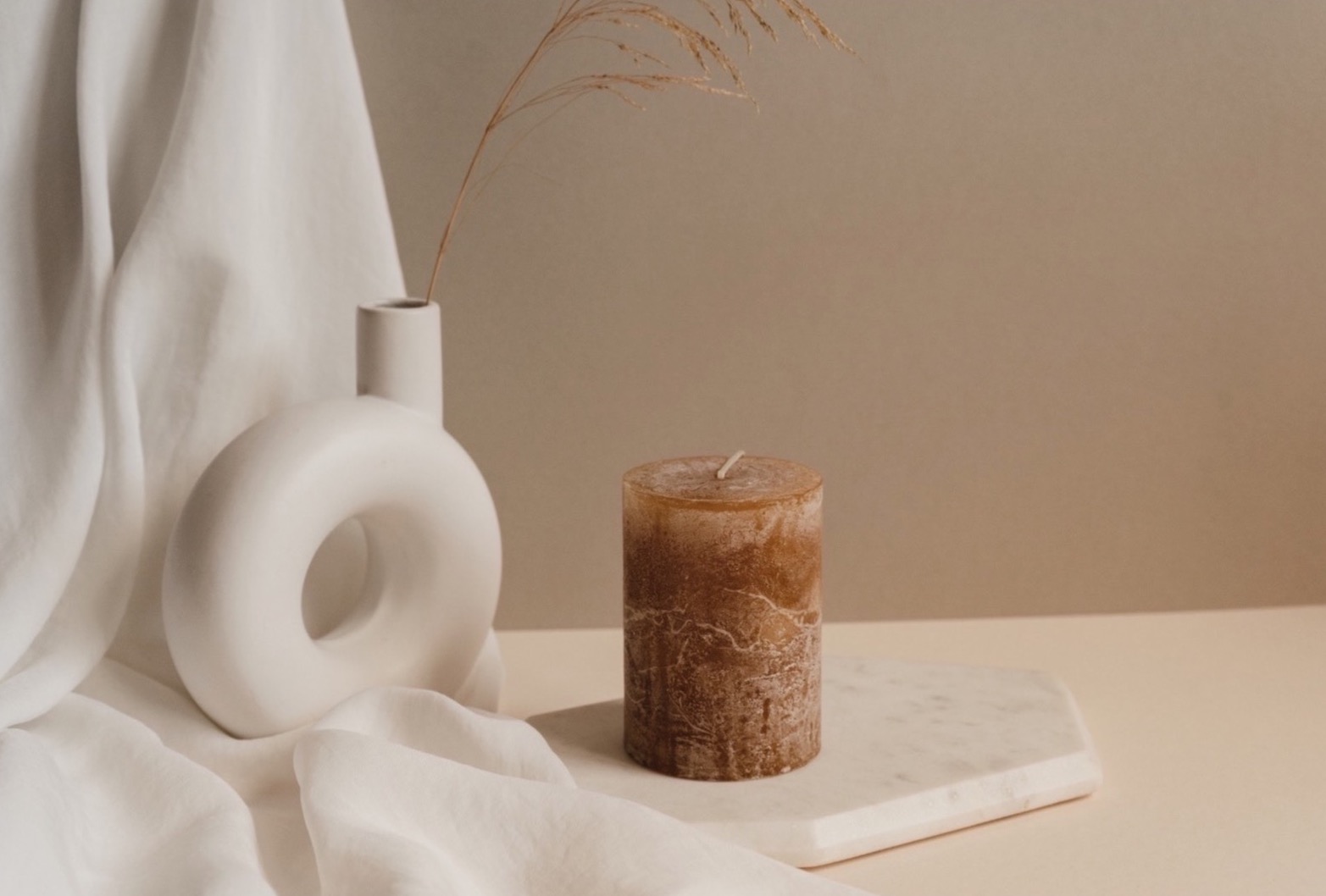 Brązowa, cylindryczna świeca z woskowym wzorem na marmurowej podstawce, obok nowoczesny, biały wazon-okrąg z suchą trawą i drapowana biała tkanina w tle.