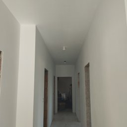 Comfort Bud - Korytarz w trakcie odświeżania, ściany pomalowane na biało, widoczne surowe obramowania drzwi i fragmenty instalacji oświetleniowej na suficie.