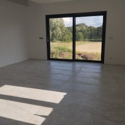 Comfort Bud - Wnętrze pustego pokoju z nową, jasną, marmurkową podłogą i dużym przesuwnym oknem balkonowym z widokiem na zewnątrz, gdzie widać trawę i drzewa.