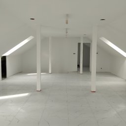 Comfort Bud - Przestronne, jasne poddasze z białymi ścianami, marmurową posadzką i filarami, przygotowane do dalszych prac wykończeniowych, widoczne otwory na oświetlenie sufitowe.