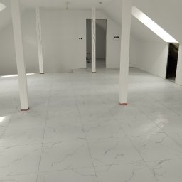 Comfort Bud - Wnętrze poddasza z białymi ścianami i podłogą wyłożoną jasnymi, marmurkowymi płytkami. Widoczne białe słupy podtrzymujące konstrukcję oraz otwory drzwiowe w ścianach.