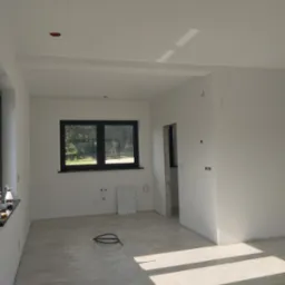 Jasne wnętrze w trakcie wykończenia: świeżo pomalowane białe ściany, okno z czarną ramą, marmurowe płytki podłogowe, widoczne instalacje elektryczne i otwór drzwiowy.