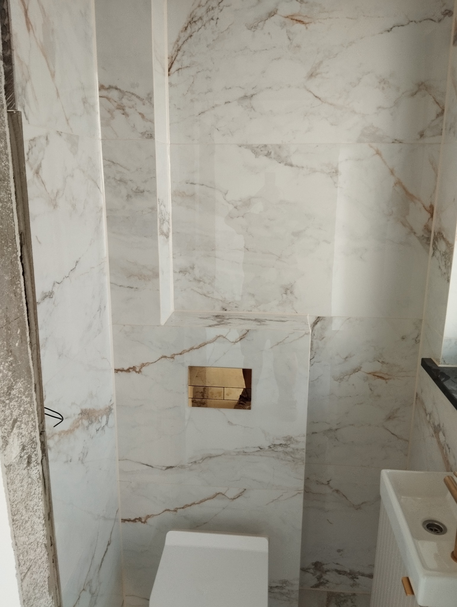 Nowoczesna łazienka z marmurowymi płytkami w odcieniach bieli i brązu, z ukrytym systemem spłukiwania toalety za złotą płytką i minimalistyczną umywalką.