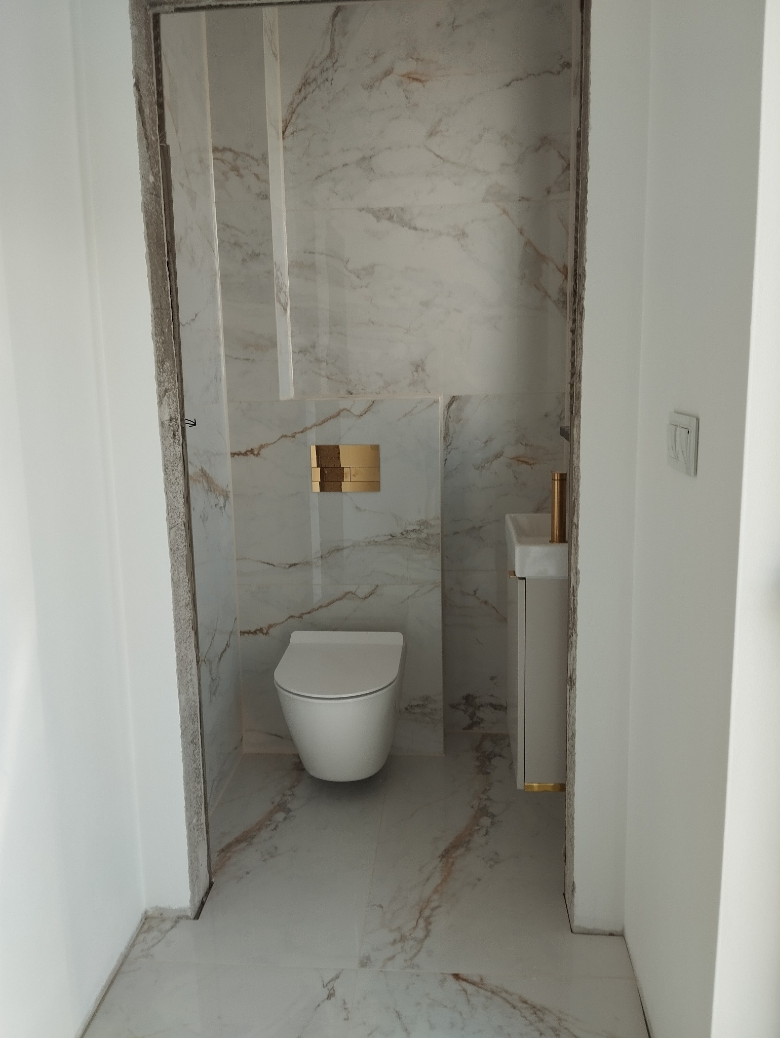 Nowoczesna łazienka w trakcie wykończenia, widoczna podwieszana toaleta, umywalka na szafce i marmurowe płytki na ścianach, surowy stan wykończenia wokół ościeżnicy.