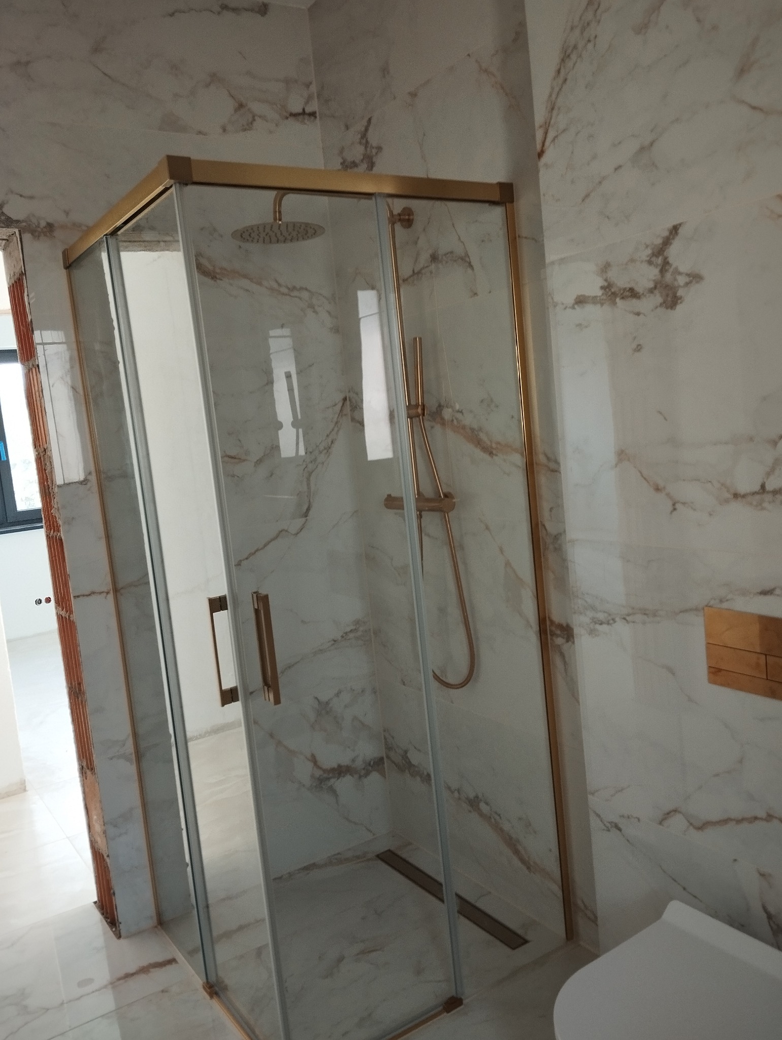 Nowoczesna kabina prysznicowa z elementami w kolorze złotym, marmurowe płytki na ścianach i podłodze, widoczny odpływ liniowy, częściowo wykończona ściana z cegły w tle.